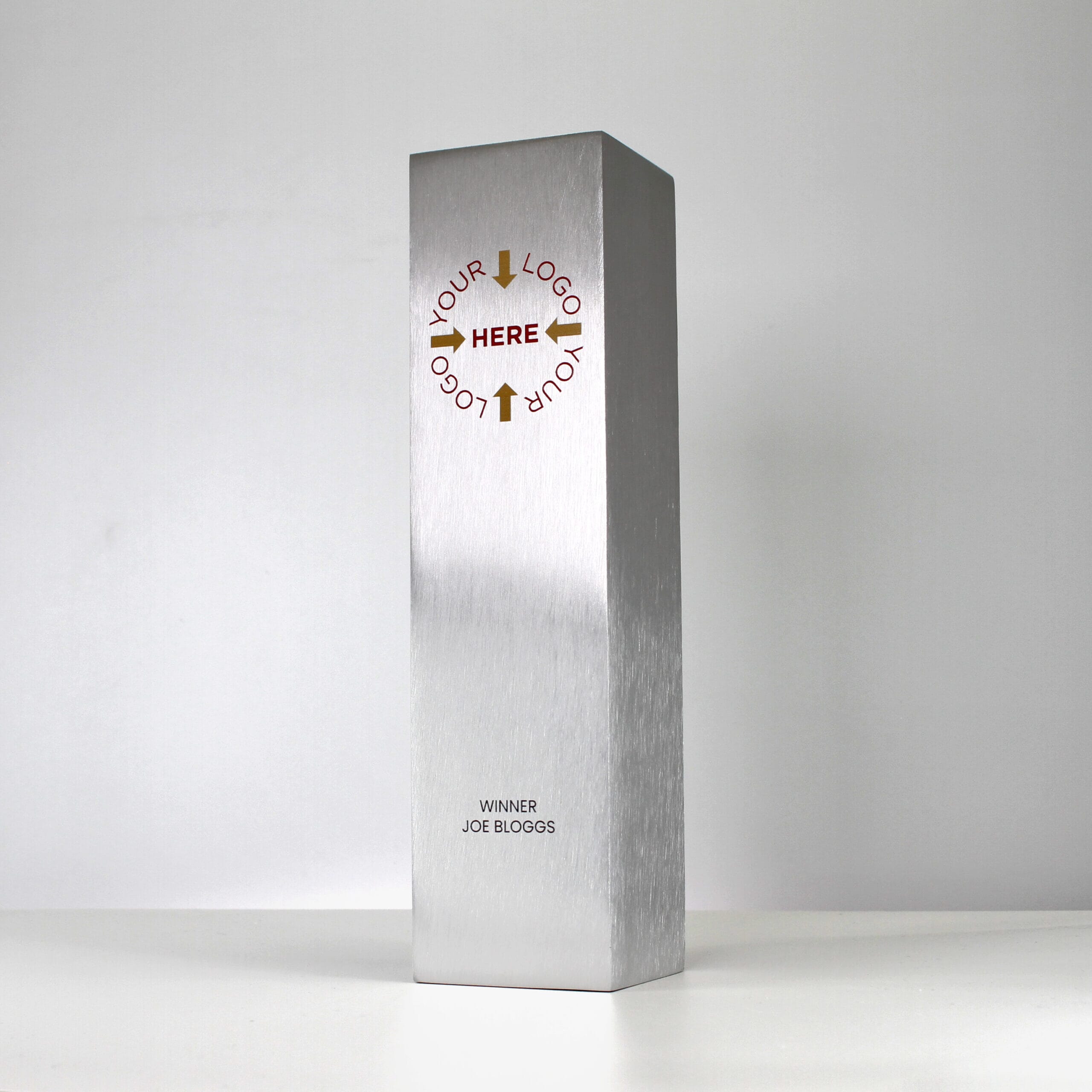 A large custom metal trophy in a column shape with 'Your Logo Here' printed in gold and red, and 'Winner Joe Bloggs' printed in black