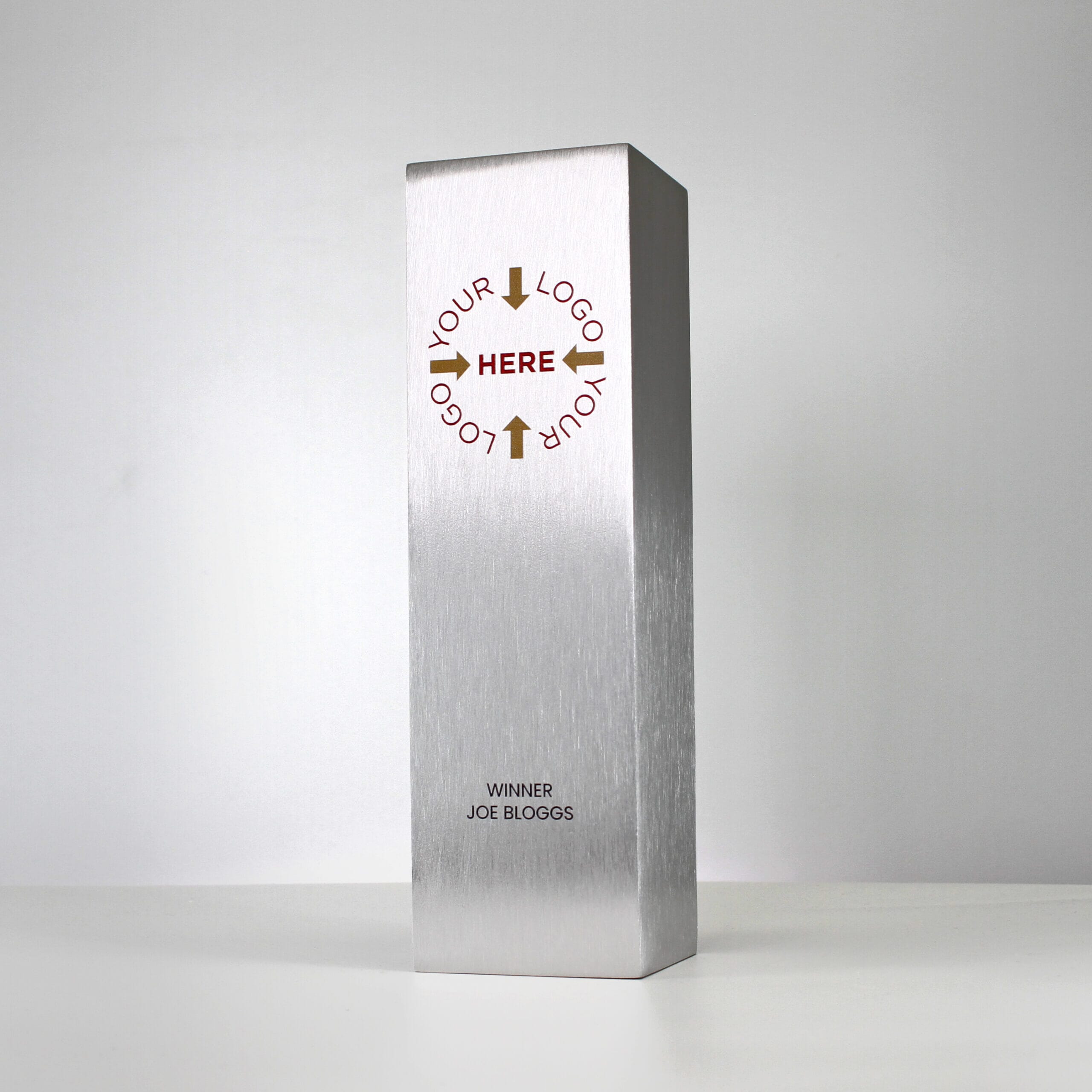 A small custom metal trophy in a column shape with 'Your Logo Here' printed in gold and red, and 'Winner Joe Bloggs' printed in black